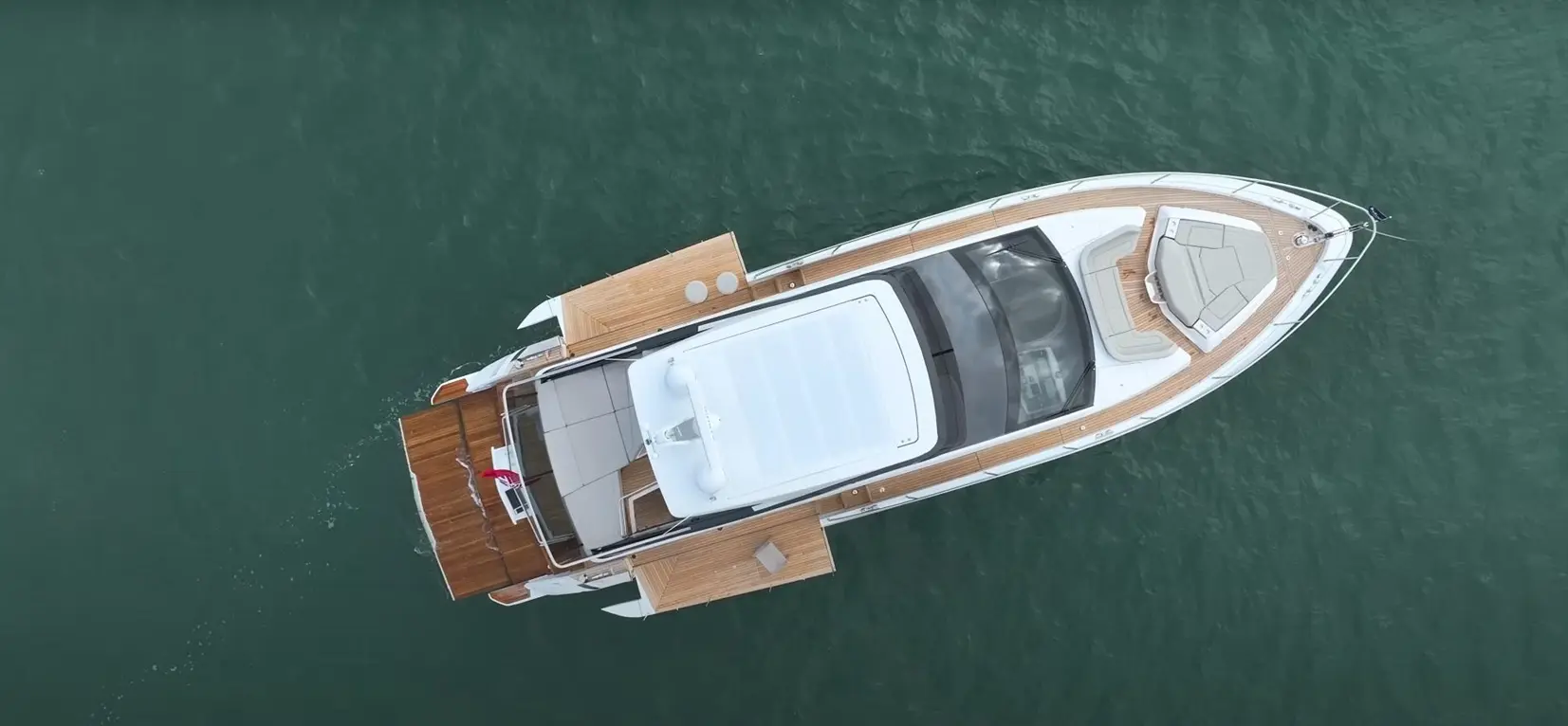 An aerial view of an elegant yacht with wooden decking and a white hull floating on green water.